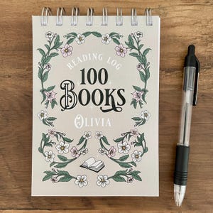 May include: A spiral-bound reading log notebook with a light grey cover featuring a floral border, the text Reading Log 100 Books, and the name Olivia in white. A black retractable ballpoint pen is positioned to the right of the notebook.