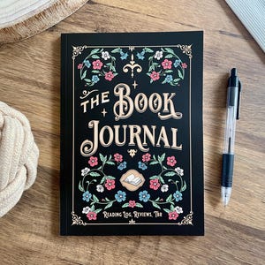 May include: A black rectangular book journal with gold lettering that reads The Book Journal and Reading Log, Reviews, TBR. The cover features a decorative floral border with pink and blue flowers and a small central icon of an open book.
