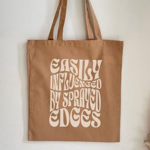 Pode incluir: Uma bolsa de lona marrom com texto branco que diz "EASILY INFLUENCED BY SPRAYED EDGES".