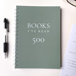 May include: A spiral-bound notebook with a sage green cover. The cover reads "BOOKS I'VE READ" above the number "500" in white text. A black pen rests on the left side of the notebook. A page with rating stars is visible on the right.