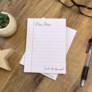 May include: White notepad with blue horizontal lines and a red vertical margin line. The notepad has the text "Miss Johnson", "Small notes, big impact", and "Reminders" in black script. A pen, glasses, and a small plant are in the background.