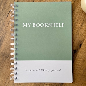 May include: A spiral-bound notebook with a sage green cover and the words "MY BOOKSHELF" in white. The bottom section is white with the text "a personal library journal". The notebook is open on a wooden surface.