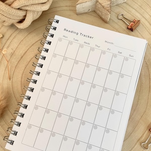 Puede incluir: Un cuaderno blanco con espiral para seguimiento de lectura con un dise&ntilde;o de cuadr&iacute;cula para entradas diarias. El cuaderno presenta el texto "Reading Tracker" en la parte superior, con espacios para los d&iacute;as de la semana y un mes. El cuaderno est&aacute; abierto, revelando p&aacute;ginas en blanco.