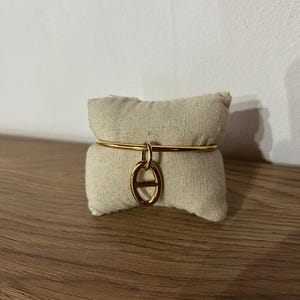 Peut inclure: Un bracelet jonc en métal doré composé d'un anneau circulaire fin et lisse, agrémenté d'un pendentif central en forme de maillon ovale avec une barre horizontale au milieu, présenté autour d'un petit coussin rectangulaire en tissu beige.
