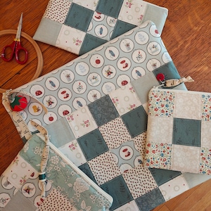 May include: A collection of handmade quilted pouches and a small sewing mat. The pouches feature patchwork designs with floral and geometric patterns in shades of blue, cream, and red. A pair of red scissors and a wooden embroidery hoop are also visible.