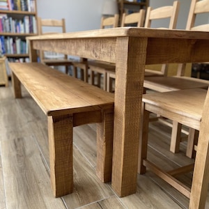 May include: A long wooden table with a matching bench and chairs. The table and chairs are made of natural wood with a rustic finish. The table is set up in a dining room with a bookshelf in the background.