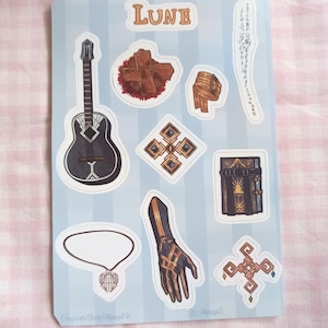 Op de afbeelding: Een stickervel met diverse illustraties met een fantasiethema. Bevat een zwarte gitaar, een boek, een handschoen en andere decoratieve elementen. Het woord "LUNE" staat bovenaan. De achtergrond is lichtblauw met verticale strepen.