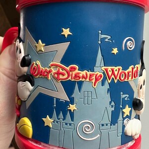Może przedstawiać: Niebieski ceramiczny kubek z czerwonym obramowaniem i uchwytem, z napisem "Walt Disney World" w kolorze czerwonym i ż&oacute;łtym. Kubek ma wz&oacute;r zamku i postacie Myszki Miki po bokach, z ż&oacute;łtymi gwiazdkami.
