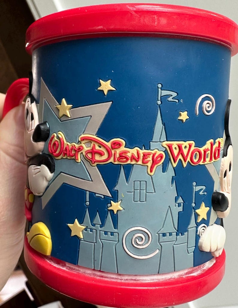 Może przedstawiać: Niebieski ceramiczny kubek z czerwonym obramowaniem i uchwytem, z napisem "Walt Disney World" w kolorze czerwonym i ż&oacute;łtym. Kubek ma wz&oacute;r zamku i postacie Myszki Miki po bokach, z ż&oacute;łtymi gwiazdkami.