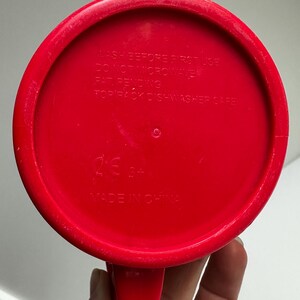 Może przedstawiać: Czerwony plastikowy kubek z uchwytem, ​​widziany od dołu. Na spodzie kubka znajduje się napis "WASH BEFORE FIRST USE", "DO NOT MICROWAVE", "PAT PENDING" i "TOP RACK DISHWASHER SAFE". Kubek jest oznaczony napisem "MADE IN CHINA".