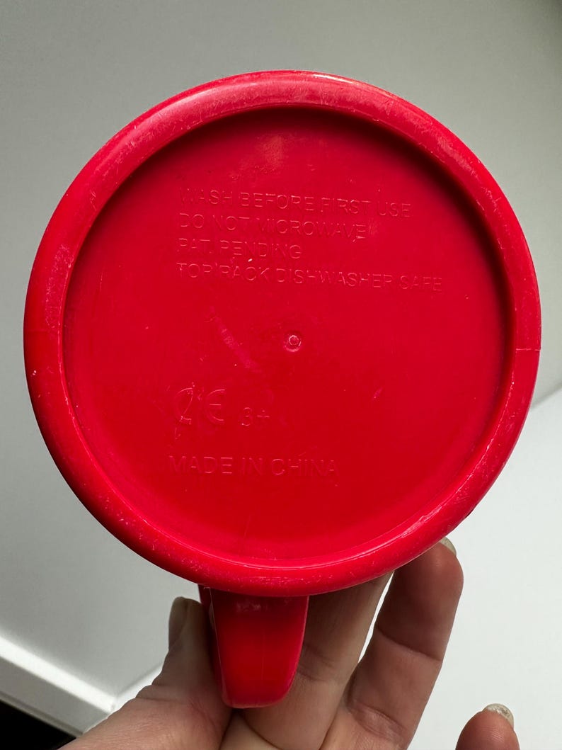 Może przedstawiać: Czerwony plastikowy kubek z uchwytem, ​​widziany od dołu. Na spodzie kubka znajduje się napis "WASH BEFORE FIRST USE", "DO NOT MICROWAVE", "PAT PENDING" i "TOP RACK DISHWASHER SAFE". Kubek jest oznaczony napisem "MADE IN CHINA".
