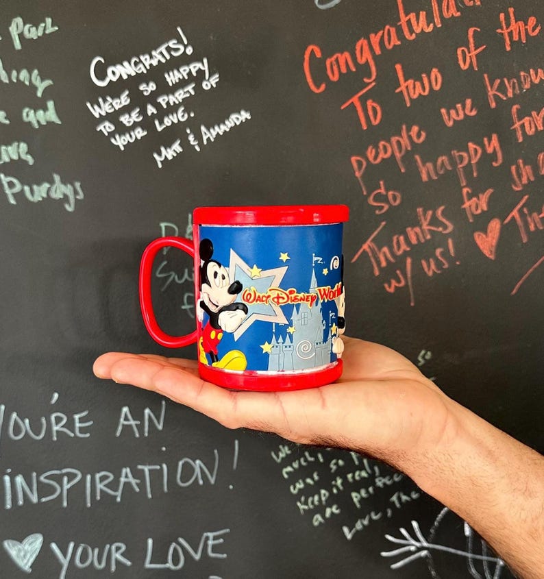Może przedstawiać: Niebiesko-czerwony kubek z Disney World z Myszka Miki i logo parku. Kubek jest trzymany w dłoni na tle tablicy z odręcznymi wiadomościami.