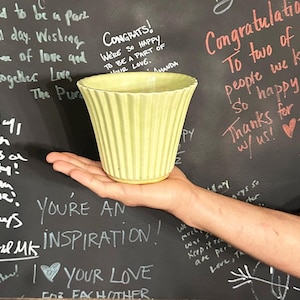 May include: A pale yellow, ribbed ceramic vase is held in a hand. The vase has a flared top and a rounded base. The background is a dark chalkboard with white and red handwritten messages, including "Congratulations!" and "You're an inspiration!"