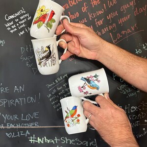 May include: Four white ceramic mugs with bird illustrations are held against a dark background with handwritten notes. Each mug features a different colorful bird design, including a cardinal, a bluebird, and a painted bunting. The mugs have a classic handle design.