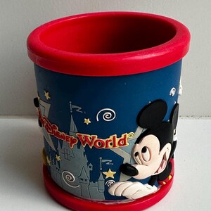 Może przedstawiać: Cylindryczny kubek pamiątkowy z Disney World. Kubek jest gł&oacute;wnie niebieski z czerwonym obramowaniem i podstawą. Myszka Miki znajduje się z boku, wraz ze słowami "Disney World" i grafiką zamku. Wykonany z twardego plastiku.