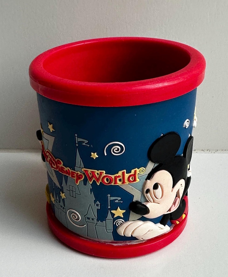 Może przedstawiać: Cylindryczny kubek pamiątkowy z Disney World. Kubek jest gł&oacute;wnie niebieski z czerwonym obramowaniem i podstawą. Myszka Miki znajduje się z boku, wraz ze słowami "Disney World" i grafiką zamku. Wykonany z twardego plastiku.