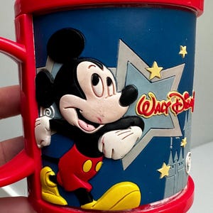 Może przedstawiać: Czerwono-niebieski kubek z Myszka Miki z tr&oacute;jwymiarowym wzorem. Kubek przedstawia Myszkę Miki w klasycznym stroju, z napisem "Walt Disney" z boku. Kubek ma czerwony uchwyt i obręcz.