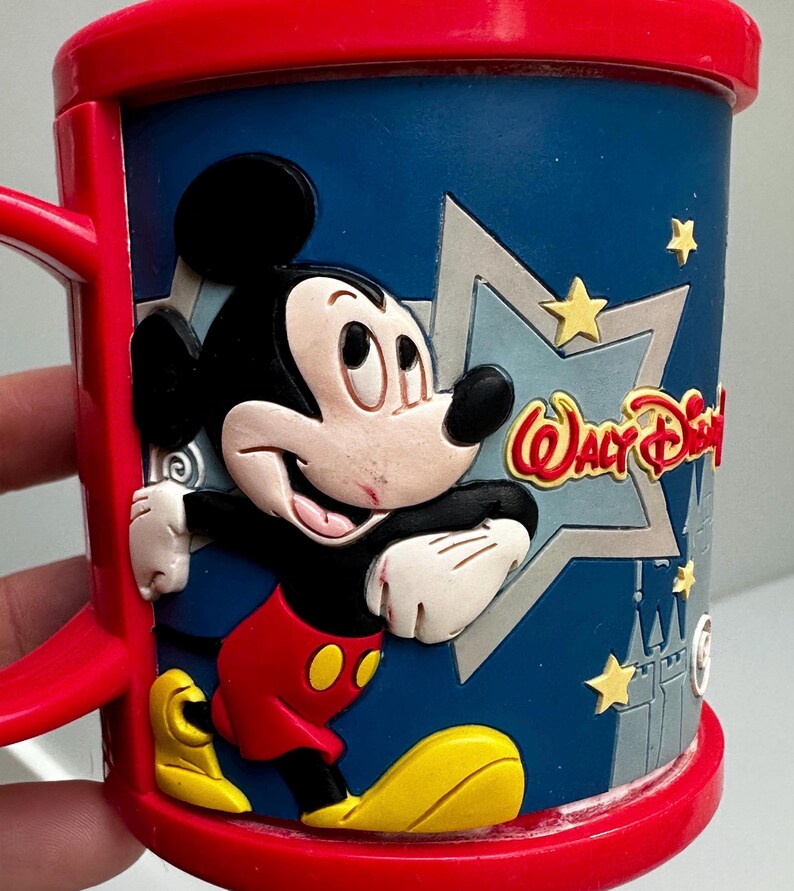Może przedstawiać: Czerwono-niebieski kubek z Myszka Miki z tr&oacute;jwymiarowym wzorem. Kubek przedstawia Myszkę Miki w klasycznym stroju, z napisem "Walt Disney" z boku. Kubek ma czerwony uchwyt i obręcz.