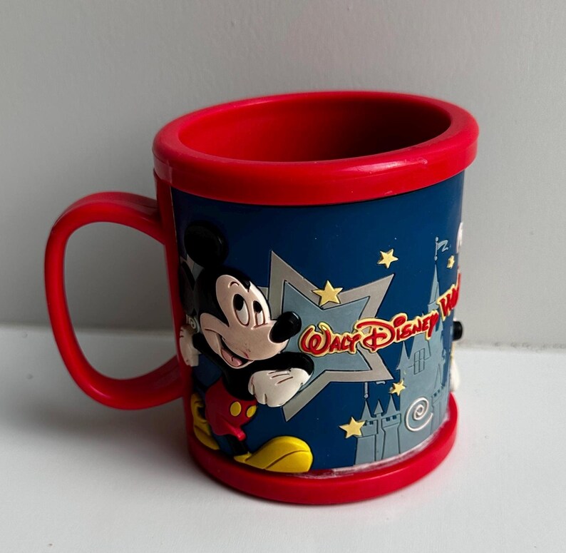 Może przedstawiać: Czerwony kubek z uchwytem i niebieskim korpusem z Myszka Miki i napisem "Walt Disney World". Kubek ma czerwoną obręcz i podstawę na białym tle.