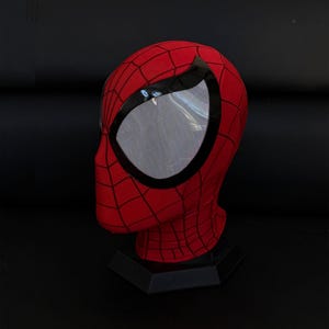 May include: A red Spider-Man mask with black webbing and large, white mesh eye openings. The mask is displayed on a black stand against a dark background. The mask is a full head covering.