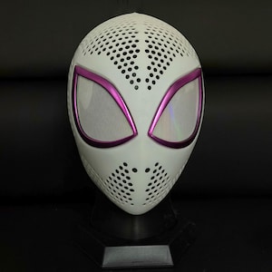 May include: A white mask with a perforated design, featuring large eye openings outlined in bright pink. The eye openings have a mesh screen. The mask is displayed on a black stand.