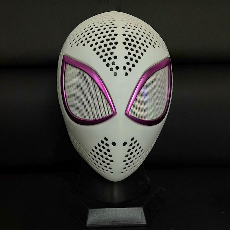May include: A white mask with a perforated design, featuring large eye openings outlined in bright pink. The eye openings have a mesh screen. The mask is displayed on a black stand.