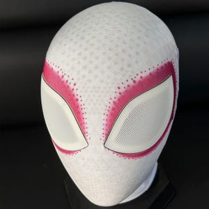 Puede incluir: Una m&aacute;scara blanca de Spider-Gwen con detalles rosas alrededor de las aberturas de los ojos. La m&aacute;scara tiene una superficie texturizada con peque&ntilde;os puntos espaciados uniformemente. Las aberturas de los ojos son blancas con un material similar a una malla.