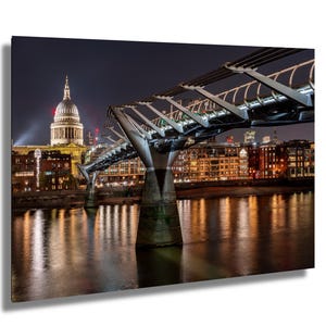 May include: A nighttime cityscape print featuring the Millennium Bridge in London, with St. Paul's Cathedral in the background. The bridge is a modern steel structure, and the buildings are illuminated with warm lights, reflecting in the water below.