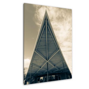 May include: Sepia-toned photograph of a modern building with a distinctive triangular roof. The roof's design features a series of parallel lines, creating a textured effect. The building's facade includes glass doors and windows, with a cloudy sky in the background.