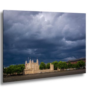 Op de afbeelding: Een foto van de Tower of London onder een dramatische, bewolkte lucht. Het historische kasteel is beige met meerdere torens en kantelen, omgeven door groene bomen en een donkere rivier. De lucht is gevuld met donkere, dreigende wolken.