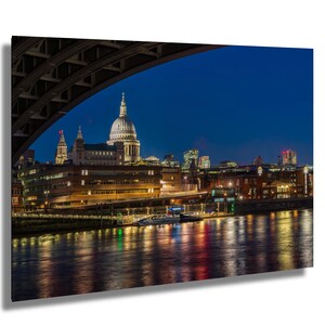 May include: A nighttime cityscape of London, featuring St. Paul's Cathedral under a dark blue sky. The image is framed by an archway, with the Thames River reflecting city lights. The scene includes buildings, bridges, and boats.