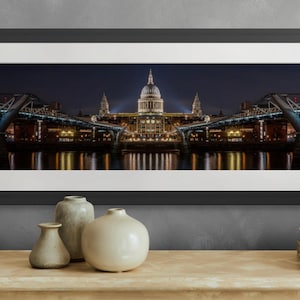 May include: A panoramic photograph of St. Paul's Cathedral in London, England at night. The cathedral is illuminated with warm yellow lights, and the reflection of the lights can be seen in the water below. The Millennium Bridge is visible in the foreground.