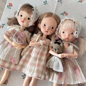 May include: Three porcelain dolls wearing dresses and hats. The dolls have blonde and brown hair. The dolls are wearing pink and white dresses with lace trim. One doll is holding a teddy bear.