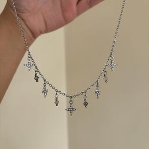 May include: Silver-toned necklace with multiple charms. The necklace features a chain with dangling charms, including crosses, lightning bolts, and crystal-like beads. The charms are adorned with small, clear crystals. The necklace is held up against a neutral background.