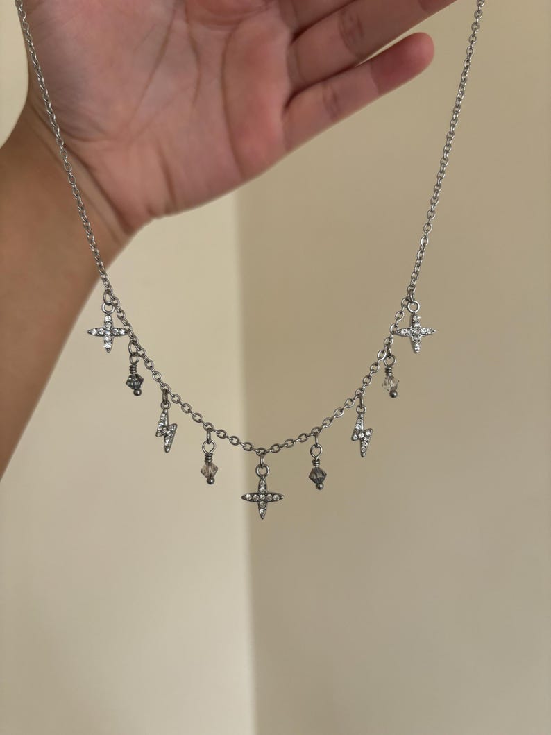 May include: Silver-toned necklace with multiple charms. The necklace features a chain with dangling charms, including crosses, lightning bolts, and crystal-like beads. The charms are adorned with small, clear crystals. The necklace is held up against a neutral background.