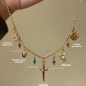 May include: A gold-coloured necklace featuring various charms: a snake, crown, acorn, leaf, and sword, embellished with colourful beads. The image includes text such as "cardan in snake form", "the cruel prince", "liriope's acorn necklace", "elfhame", "the wicked king", "the queen of nothing", "nightfell", and "cardan greenbriar".