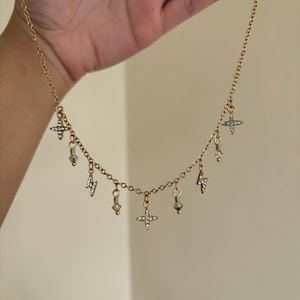 May include: A gold-colored necklace with multiple charms. The charms include small crosses, lightning bolts, and crystal-like beads. The necklace is held up by a hand against a neutral background.
