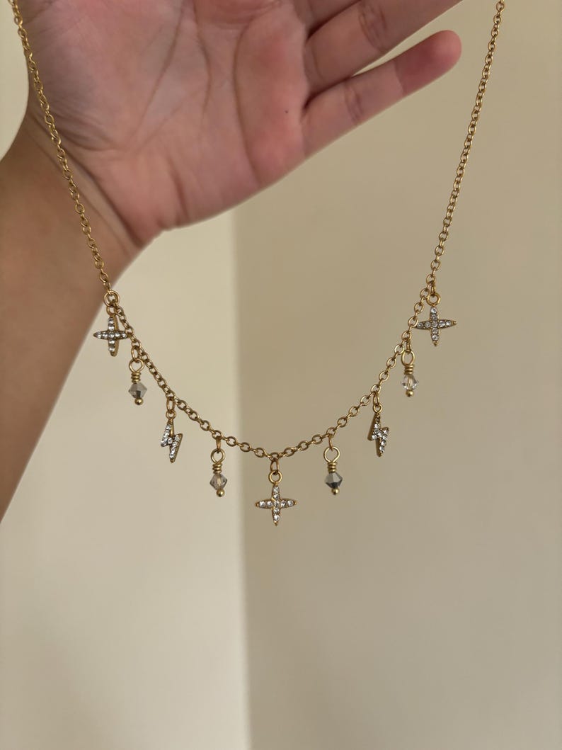 May include: A gold-colored necklace with multiple charms. The charms include small crosses, lightning bolts, and crystal-like beads. The necklace is held up by a hand against a neutral background.