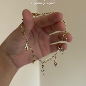 Op de afbeelding: Goudkleurige kettingarmband met bungelende bedels. De armband heeft kleine, sprankelende bliksemvormige bedels en amberkleurige kralen. De tekst "Fourth Wing - Violet's Lightning Signet" is zichtbaar.