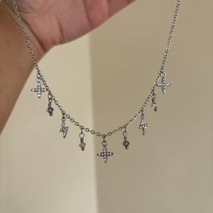 May include: Silver-toned necklace featuring a delicate chain with multiple charms. The charms include small crosses, lightning bolts, and crystal-like beads. The necklace is held up against a neutral background.