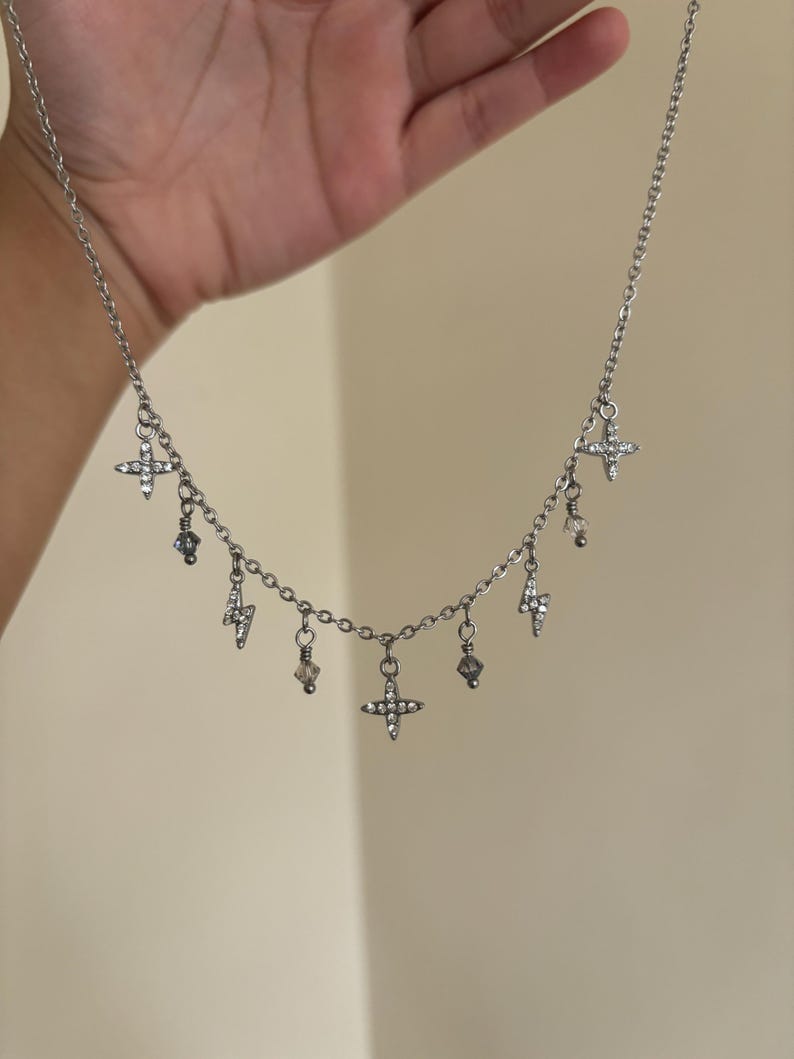 May include: Silver-toned necklace featuring a delicate chain with multiple charms. The charms include small crosses, lightning bolts, and crystal-like beads. The necklace is held up against a neutral background.