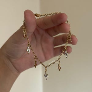 Op de afbeelding: Goudkleurige enkelband met een delicaat kettingontwerp. De enkelband is versierd met kleine, sprankelende bliksemvormige bedels en amberkleurige kralen. De sieraden worden tegen een neutrale achtergrond gehouden.