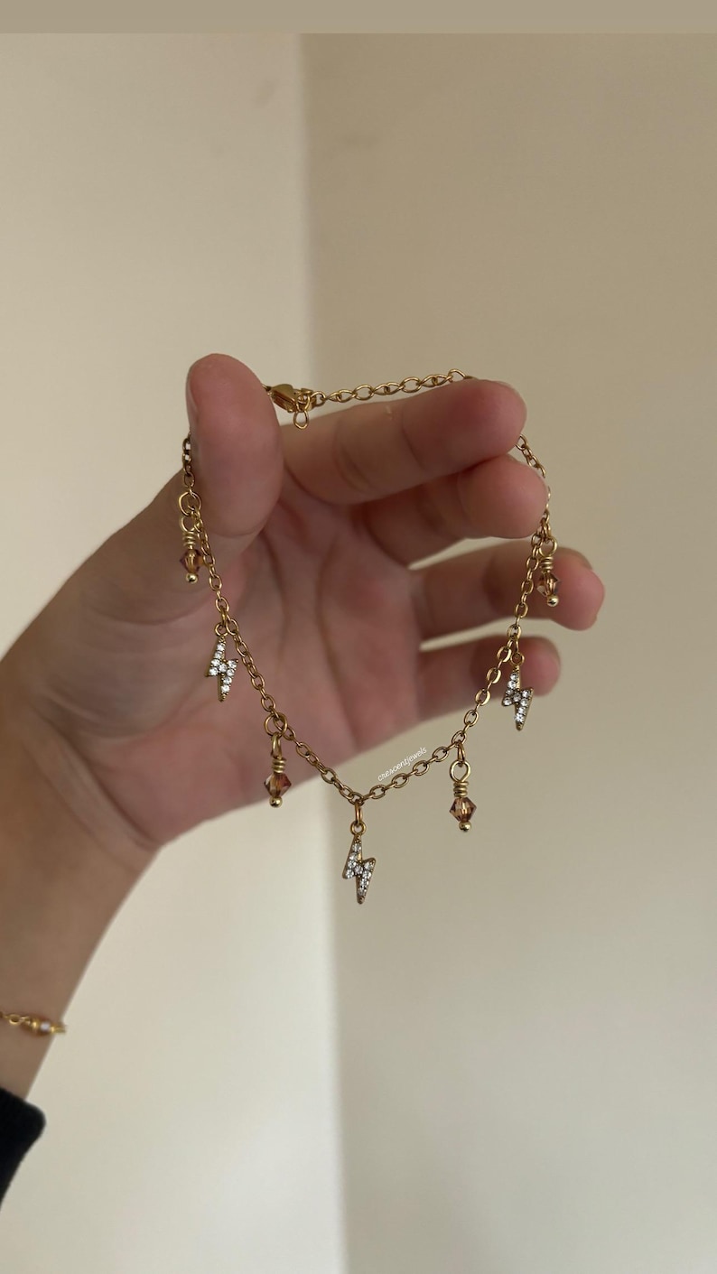 Op de afbeelding: Goudkleurige enkelband met een delicaat kettingontwerp. De enkelband is versierd met kleine, sprankelende bliksemvormige bedels en amberkleurige kralen. De sieraden worden tegen een neutrale achtergrond gehouden.