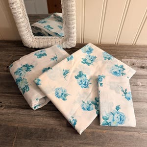 Peut inclure: Ensemble de linge de lit blanc avec un motif de roses bleues, présenté sur une surface en bois. Un miroir en osier blanc est visible en arrière-plan. L'ensemble comprend un drap plié, un drap plat et une taie d'oreiller.