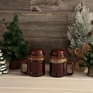 May include: Two brown ceramic mugs with lids and handles. The mugs have a glossy finish and a two-tone design, with a lighter band around the middle. The mugs are displayed on a white surface with small decorative trees in the background.