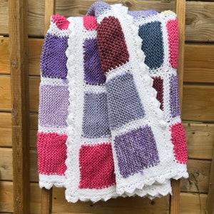 May include: A crocheted baby blanket with a white border and squares of different colours, including purple, pink, and grey.