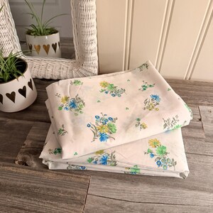 May include: White bedsheets with a floral pattern in blue, green, and yellow. The sheets are folded and stacked on a wooden surface. The floral design is repeated across the fabric. The sheets are part of a bedding set.