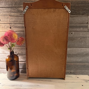 May include: A wooden wall-mounted cabinet with a light brown finish. The cabinet has a rectangular shape with a decorative top and metal brackets for hanging. A brown glass bottle holds pink and yellow flowers.