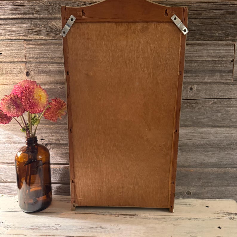 May include: A wooden wall-mounted cabinet with a light brown finish. The cabinet has a rectangular shape with a decorative top and metal brackets for hanging. A brown glass bottle holds pink and yellow flowers.