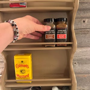 May include: A beige wooden spice rack with multiple shelves. Two glass jars of Trader Joe's spices, labeled "Ground Cumin" and "Smoked Paprika", are on a shelf. A yellow tin of Colman's Mustard is also visible. A hand is reaching for the spices.